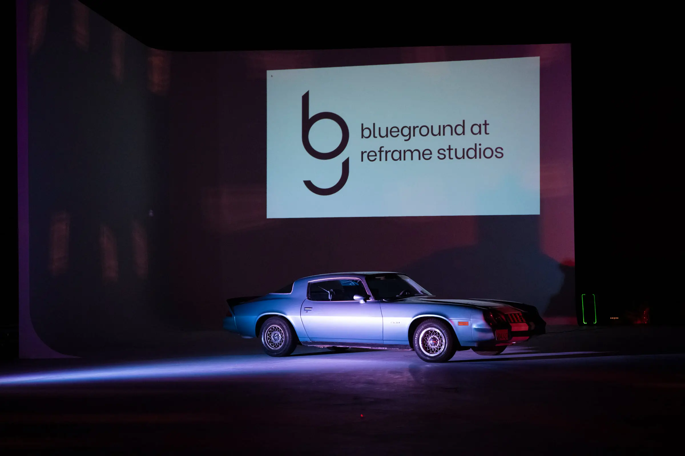 The projection screen on the studio wall showing a logo that reads blueground at reframe studios, with the blue coupe parked in profile below it under purple light and a beam of light across the floor.