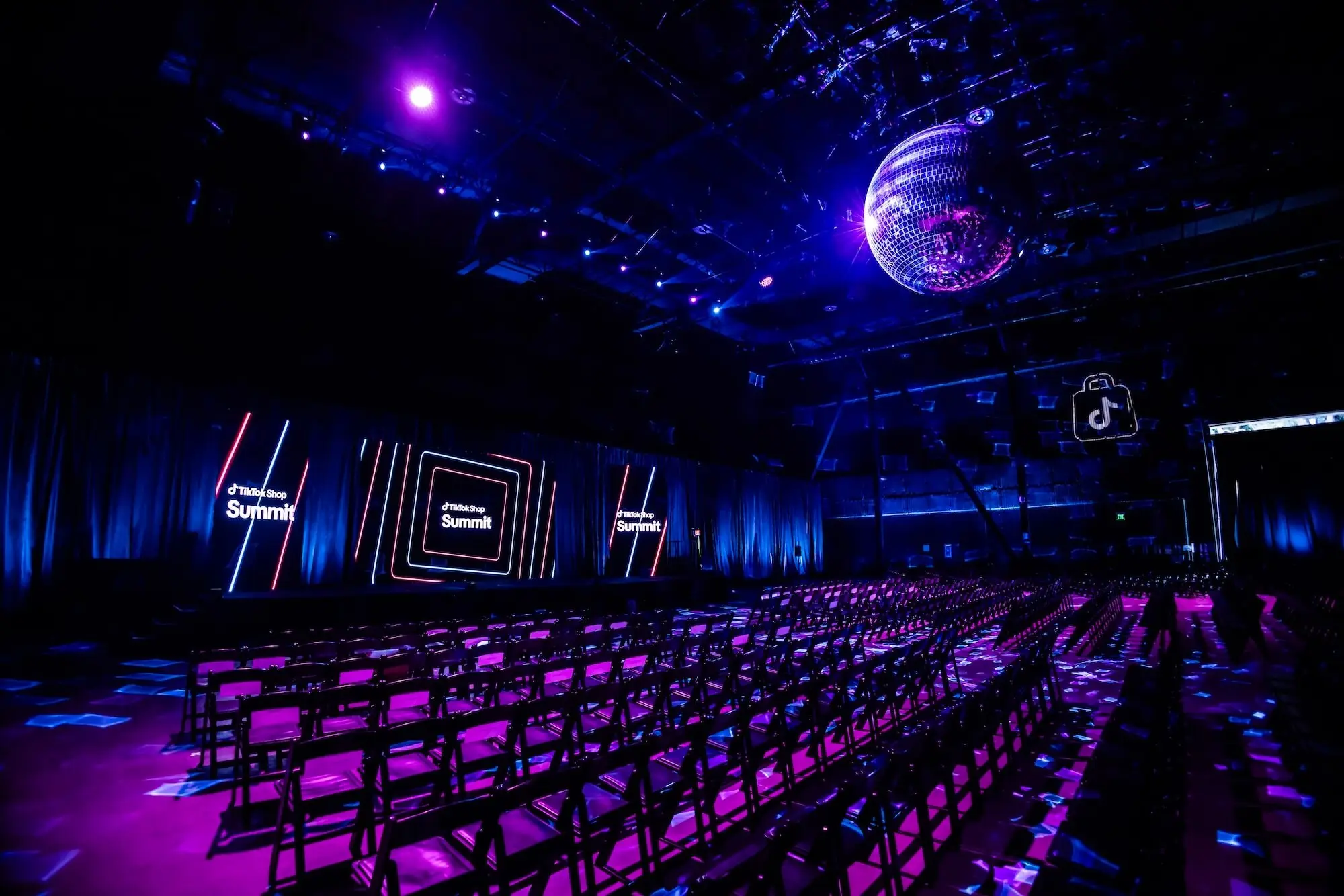 The keynote room lit magenta from the floor, Summit panels along the LED wall, a lit shop bag sign on the side wall, and the mirror ball overhead above empty chairs.