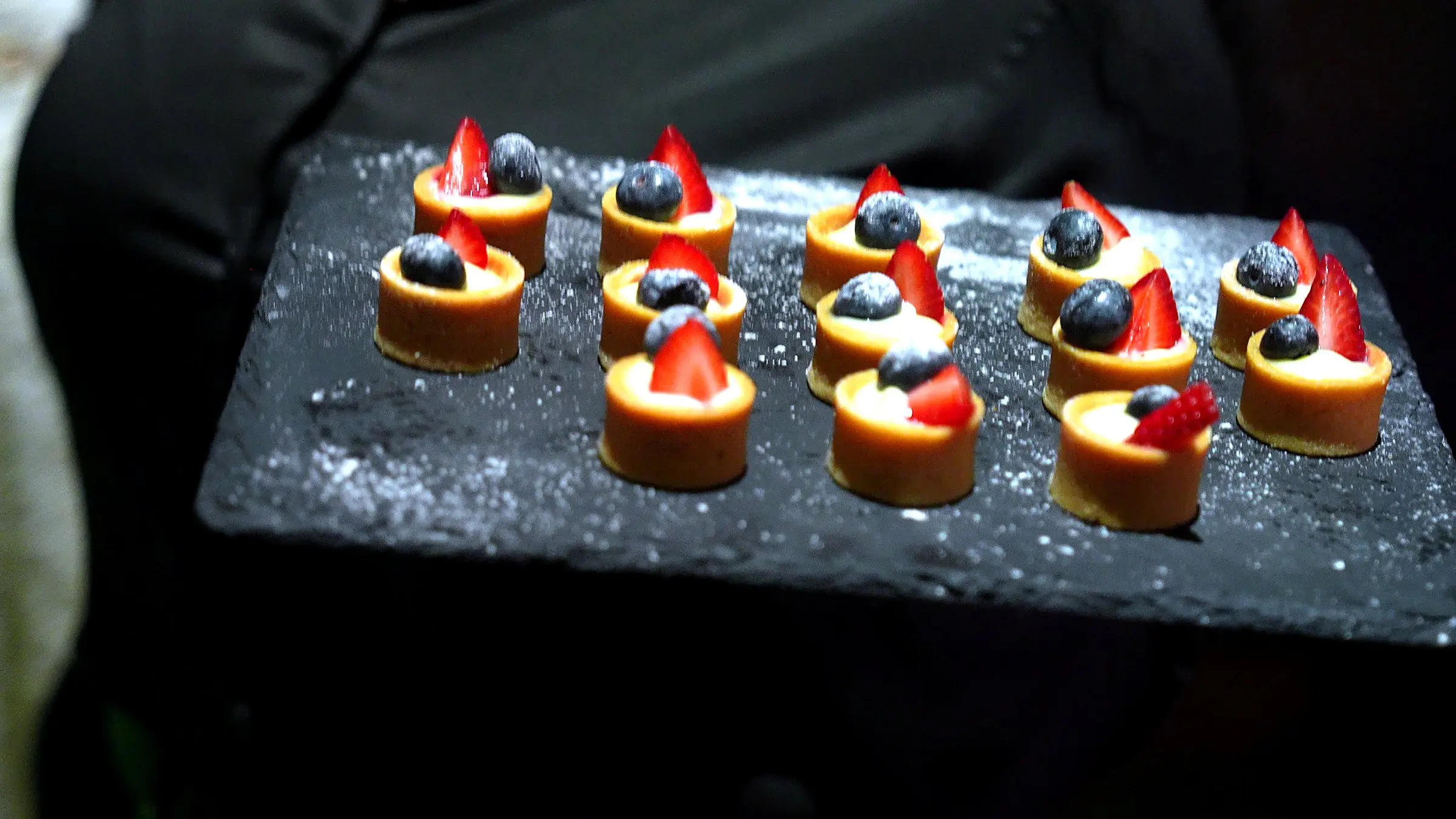 A server holding a slate tray of fruit tarts topped with strawberries and blueberries and dusted with sugar.