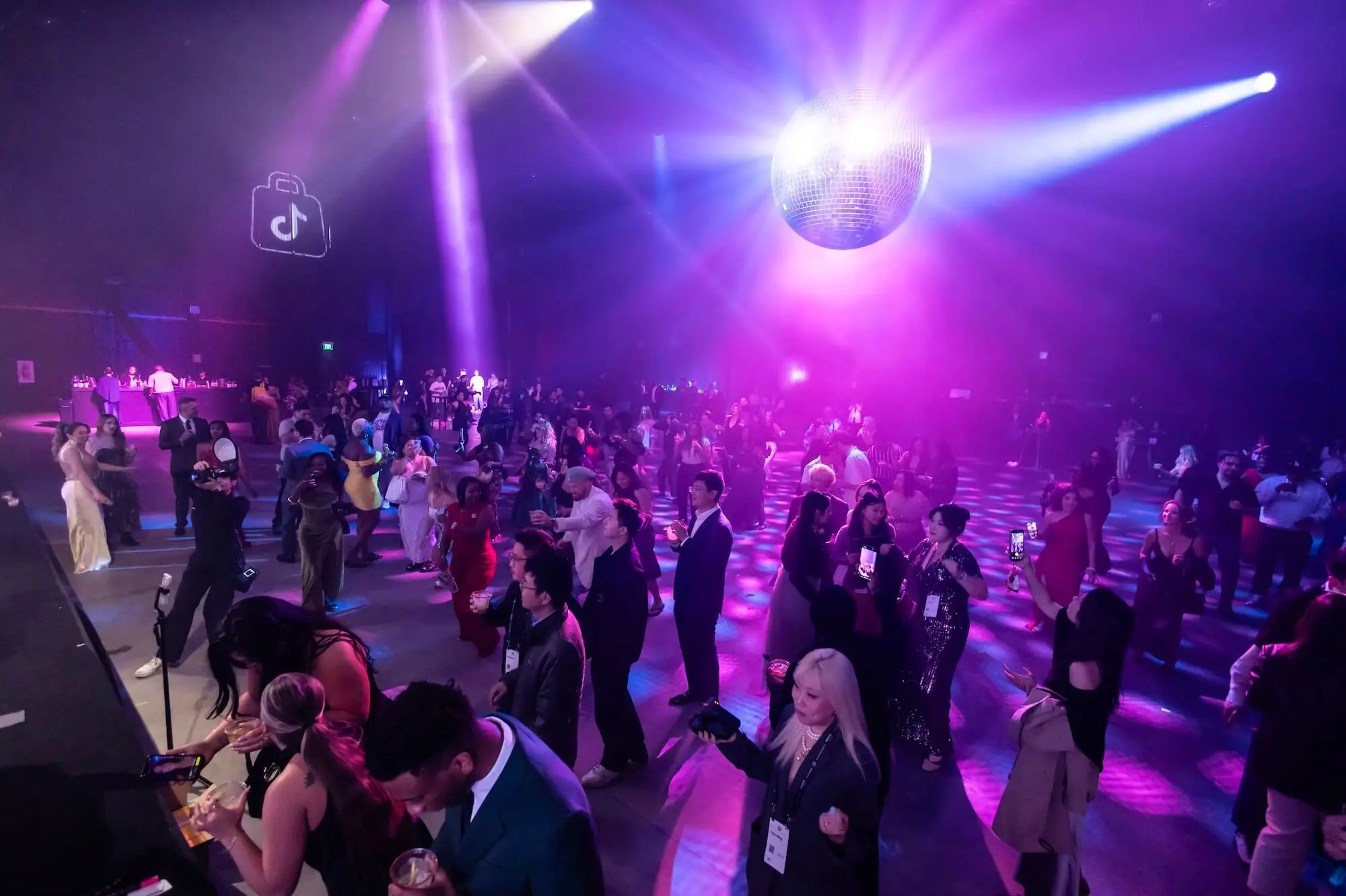 The floor packed with guests under the mirror ball, purple beams crossing the room from the overhead rig.