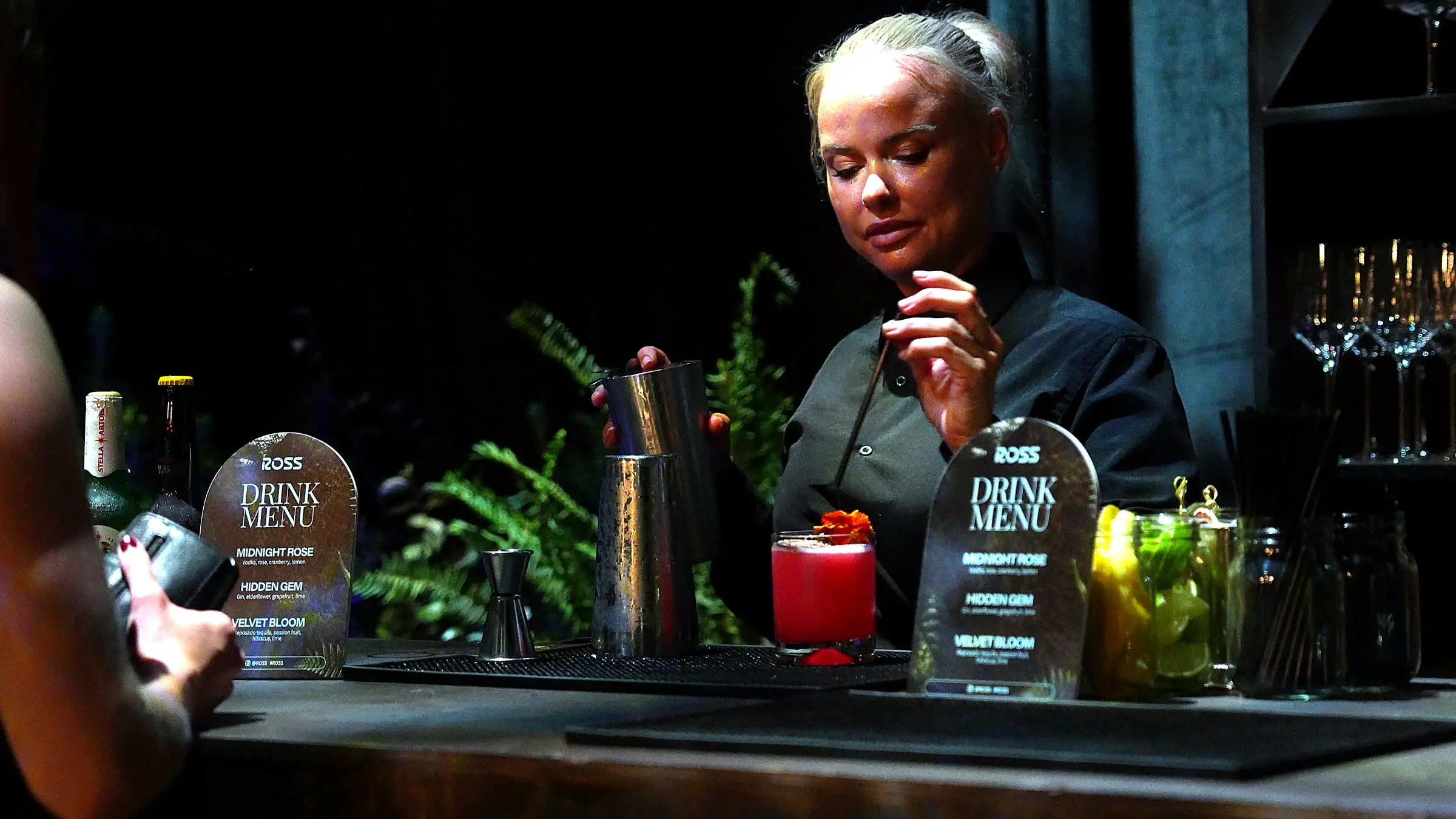 A bartender building a drink behind the bar with a shaker tin in her hand, the Ross drink menu cards and a garnished red cocktail on the mat in front of her.
