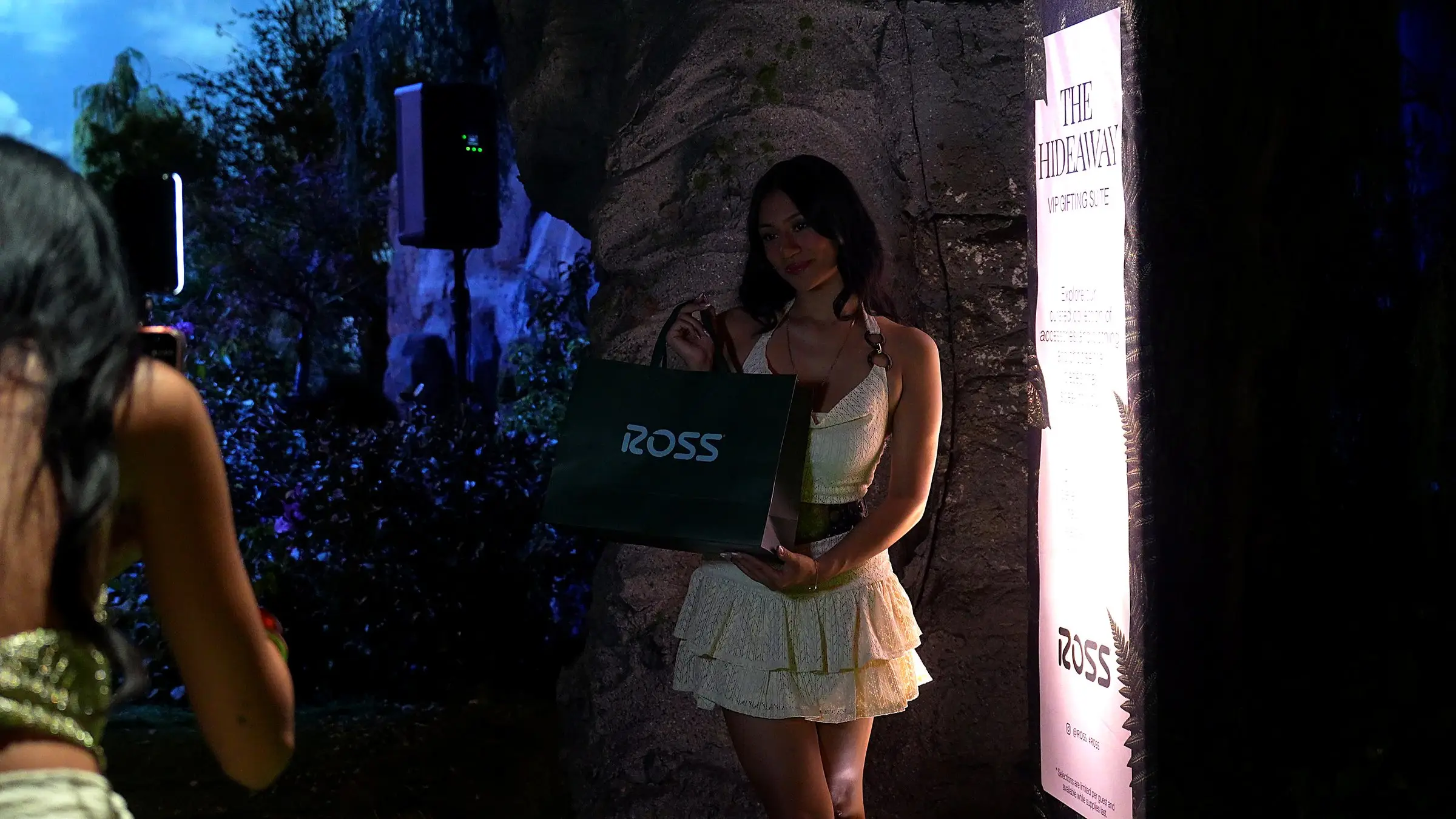A guest posing with a green Ross bag against the rock wall beside the lit sign for The Hideaway gifting suite while another guest photographs her.