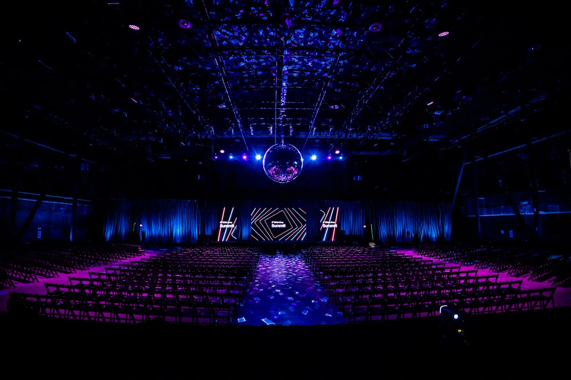 A dark arena floor lit blue, a mirror ball hung over center stage, letter set pieces along the LED wall, and rows of empty chairs under a truss grid.