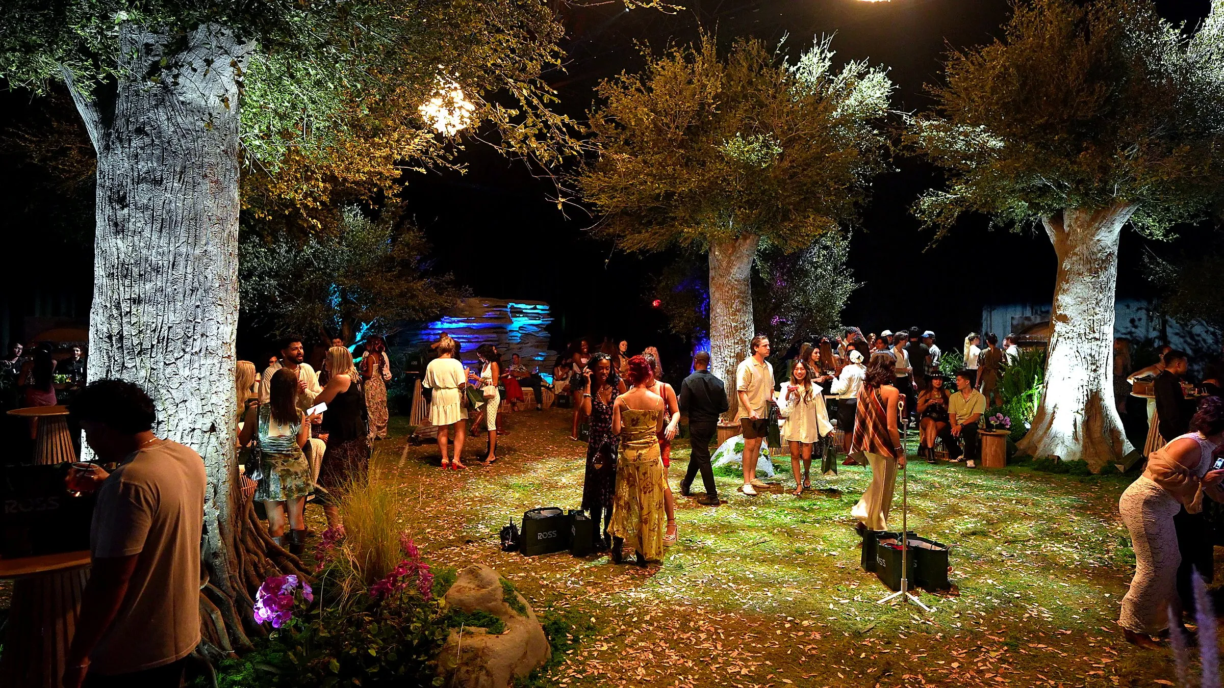 Guests spread across the leaf covered turf between three sculpted trees lit from below, a chandelier hanging in the branches above them and a stacked stone wall washed blue at the back of the room.