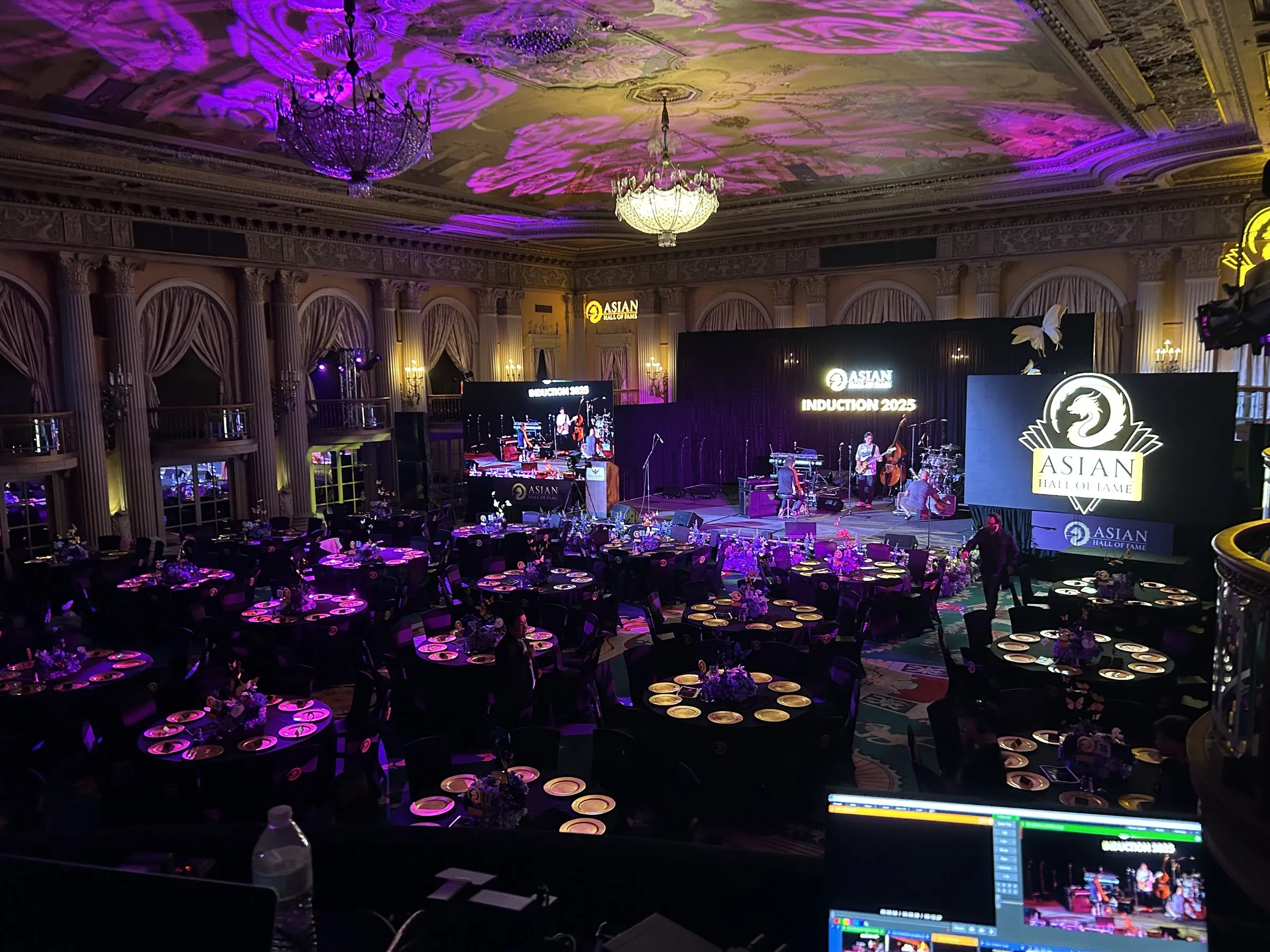A hotel ballroom washed in purple, pattern gobos across the ornate ceiling, round dinner tables filling the floor, and a lit stage with an LED screen at the far end.