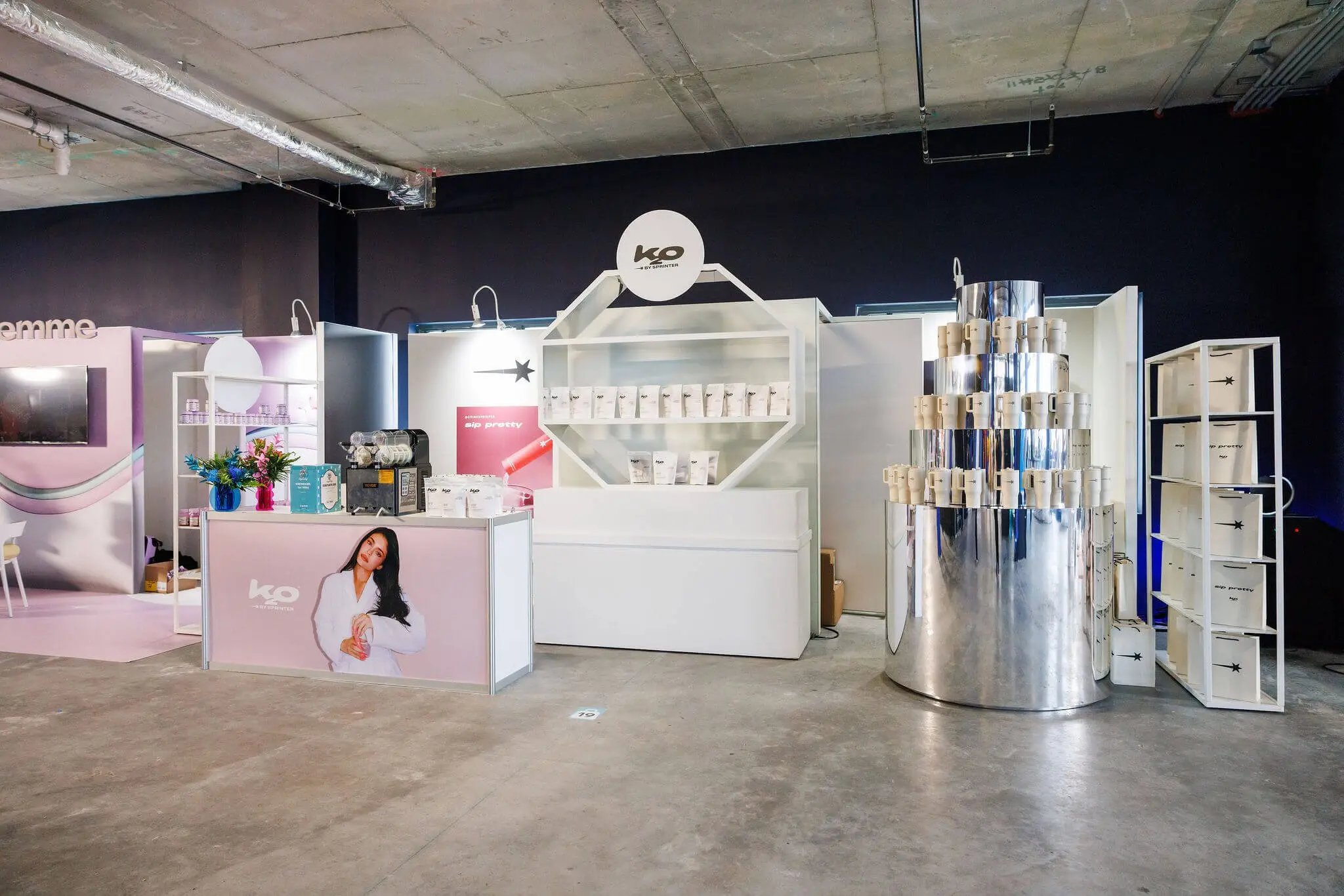 A white modular product booth with tiered shelving, a branded counter, and a mirrored column, built on a bare concrete floor.