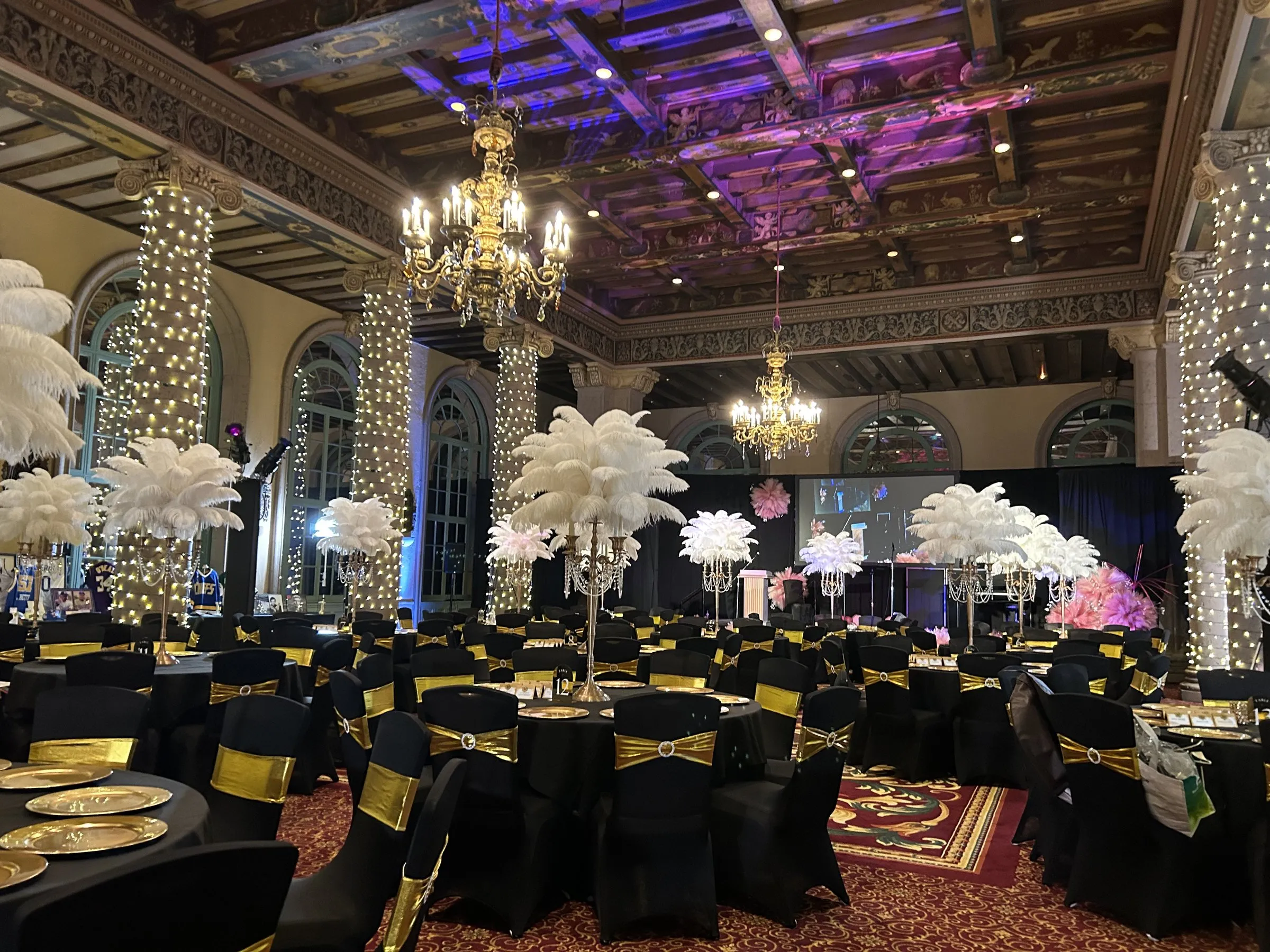 A hotel ballroom set for dinner, chandeliers and a magenta ceiling wash overhead, columns wrapped in string lights, and black tables with white feather centerpieces.