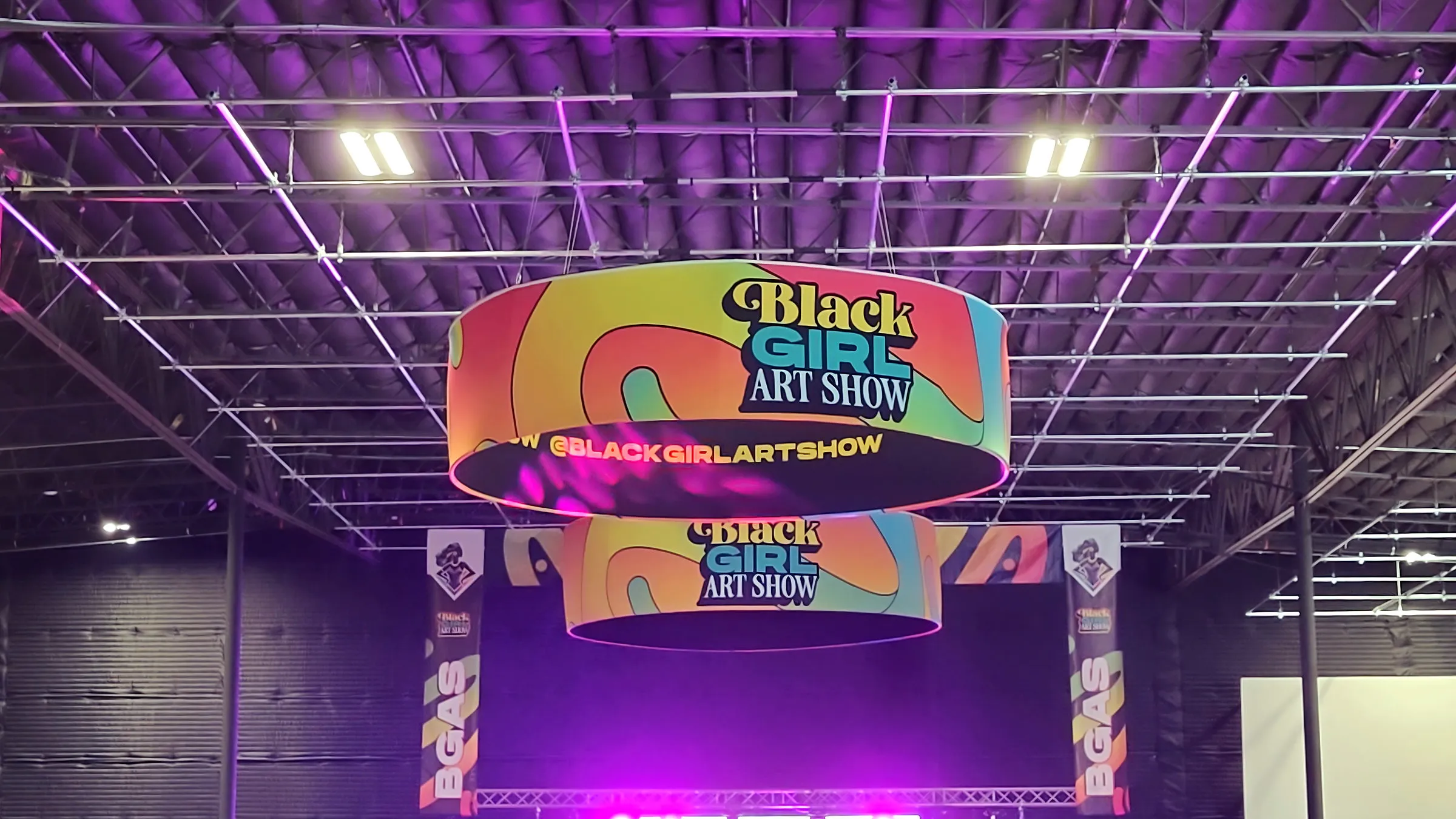 Two round Black Girl Art Show banners hung one below the other under the purple lit grid ceiling, with the stage glowing magenta on the floor below them.