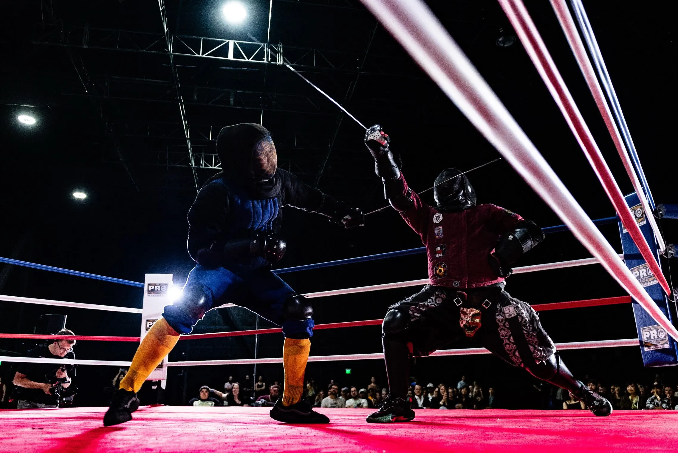 Two fighters in masks and padded jackets mid exchange on the red ring canvas, swords crossed, with the seated crowd behind the ropes and the lights overhead.