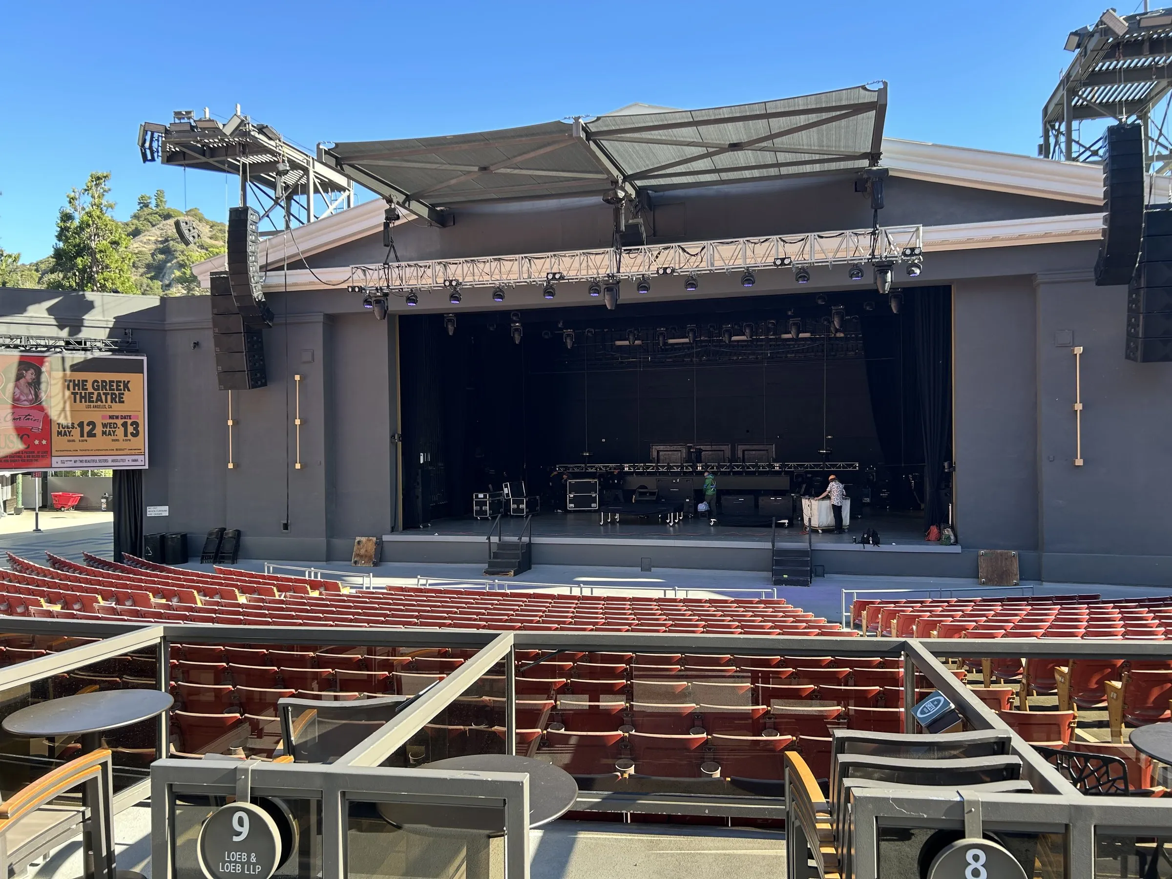 An outdoor amphitheatre stage in daylight before doors, with the roof canopy overhead, speaker arrays hung at the sides, backline set on the deck, and empty rows of seats in front.
