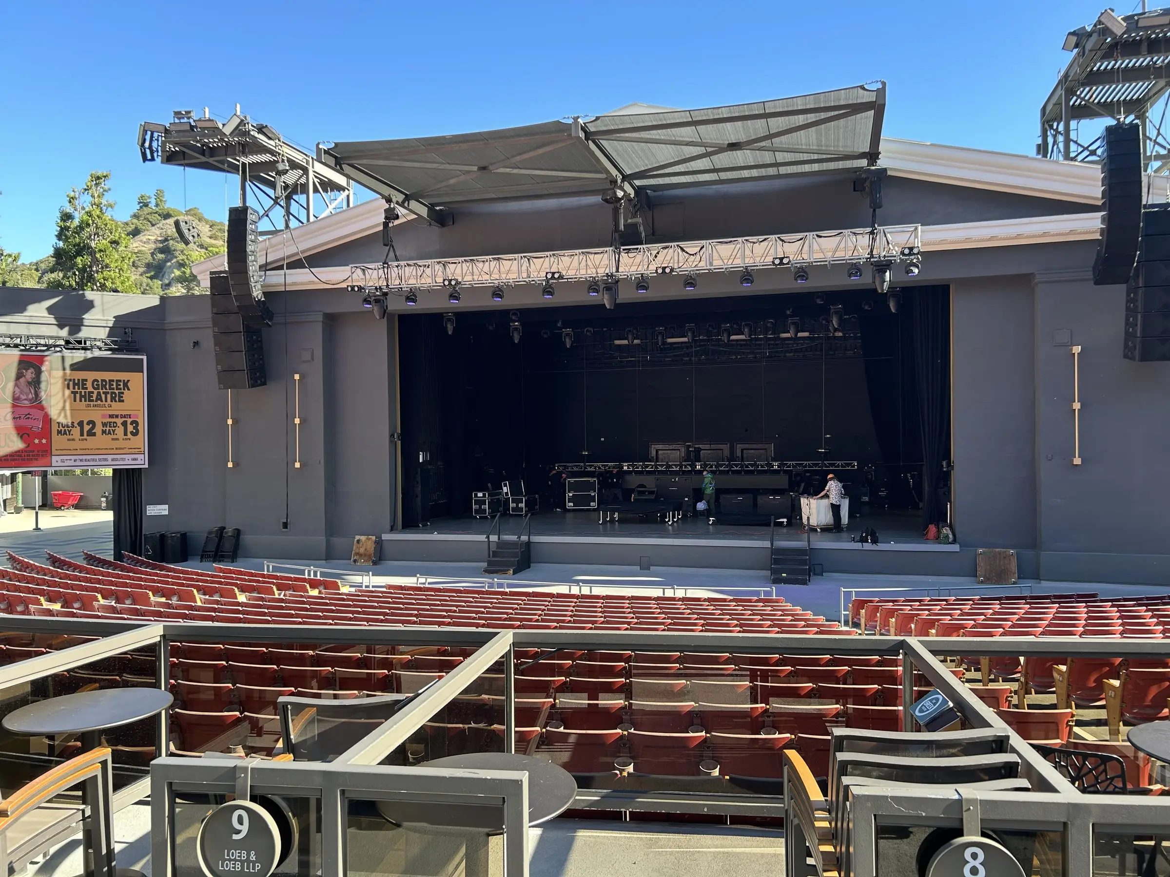 An outdoor amphitheatre stage in daylight before doors, with the roof canopy overhead, speaker arrays hung at the sides, backline set on the deck, and empty rows of seats in front.
